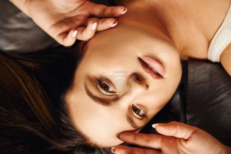 Photo for Top view of plus size woman with natural makeup lying on bed with grey bedding while touching face and looking at camera, body positive, figure type, modern apartment, portrait, close up view - Royalty Free Image