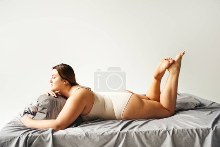 Photo for Side view of brunette and curvy woman with natural makeup wearing beige bodysuit and resting on bed with grey bedding while looking away, body positive, figure type, modern apartment - Royalty Free Image