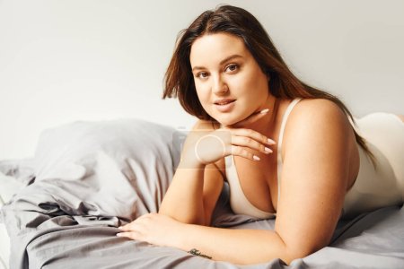 brunette plus size woman with natural makeup wearing beige bodysuit and resting on bed with grey bedding while looking at camera, body positive, figure type, tattoo translation: harmony 