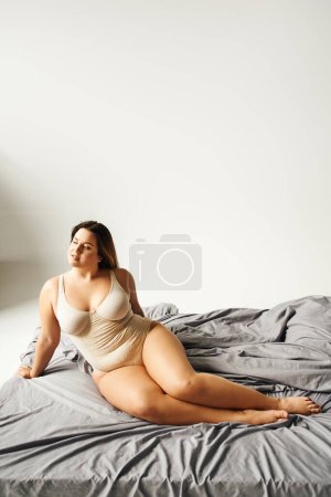 verträumte und brünette Frau mit natürlichem Make-up und Plus-Size-Körper in beigem Body und posiert auf dem Bett mit grauer Bettwäsche, körperbetont, Figurentyp, nackte Füße, wegschauen 