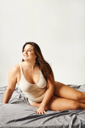 charming woman with plus size body wearing beige bodysuit and smiling and posing on bed with grey bedding, body positive, figure type, bare feet, looking away, tattoo translation: harmony