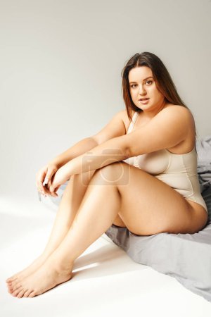 full length of charming woman with plus size body and bare feet wearing beige bodysuit and looking at camera while sitting on bed with grey bedding, body positive, figure type 
