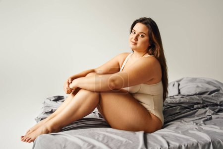 full length of brunette alluring woman with long hair and plus size body and bare feet wearing beige bodysuit and looking at camera while sitting on bed with grey bedding, body positive, figure type 