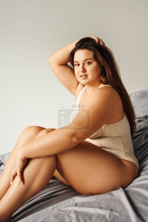 brunette alluring woman with long hair and plus size body and bare feet wearing beige bodysuit and looking at camera while touching hair and sitting on bed with grey bedding, body positive 