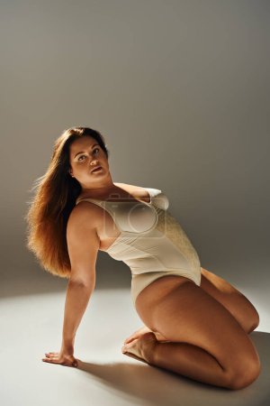 curvy woman with brunette long hair and plus size body wearing beige bodysuit and looking at camera while posing on grey background with studio light, body positive, figure type, full length 
