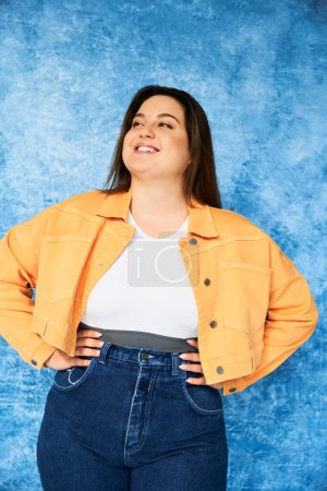 cheerful plus size woman with long hair and natural makeup wearing crop top, orange jacket and denim jeans while posing with hands on hips and looking away on mottled blue background, body positive 