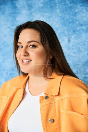 portrait of positive plus size woman with long hair and natural makeup wearing crop top and orange jacket while posing and looking at camera on mottled blue background, body positive 