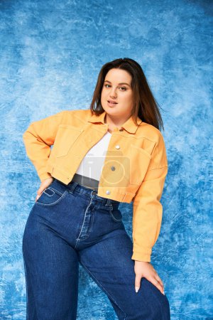 woman with plus size body and long hair, wearing crop top, orange jacket and denim jeans while posing with hand on hip and looking at camera on mottled blue background, body positive 