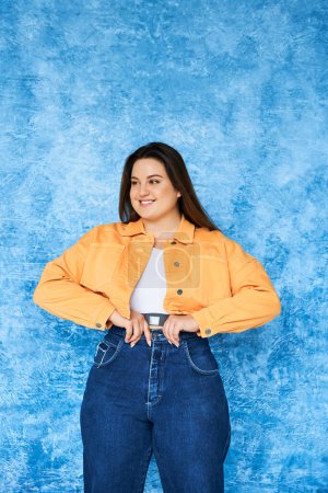 Photo for Positive plus size woman with long hair and natural makeup wearing crop top, orange jacket and adjusting denim jeans while posing and looking away on mottled blue background, body positive - Royalty Free Image