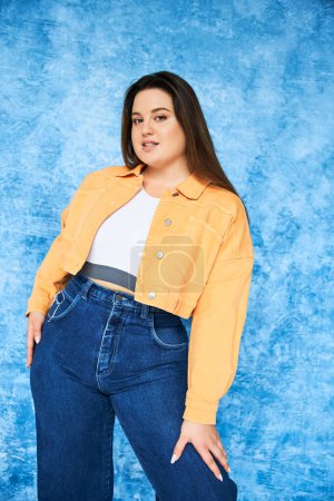 plus size woman with brunette long hair and natural makeup wearing crop top, orange jacket and denim jeans while posing and looking at camera on mottled blue background, body positive 