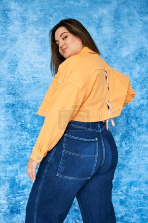 Photo for Alluring plus size woman with long hair and natural makeup wearing crop top, orange jacket and denim jeans while posing and looking at camera on mottled blue background, body positive - Royalty Free Image