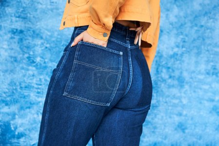 cropped view of anonymous plus size woman in orange jacket and posing with hand in pocket of denim jeans while standing on mottled blue background, body positive 