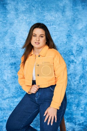 brünette plus size Frau mit langen Haaren und natürlichem Make-up trägt orangefarbene Jacke und Jeans, während sie auf einem Hocker sitzt und in die Kamera auf blau meliertem Hintergrund schaut, Körper positiv 