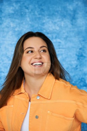 retrato de mujer feliz más tamaño con el pelo largo y maquillaje natural usando chaqueta naranja brillante y mirando hacia otro lado mientras posando sobre fondo azul moteado, cuerpo positivo 