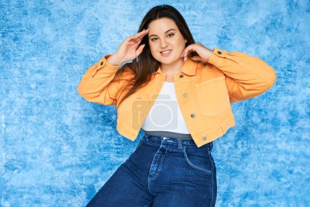 Self Acceptance, body positive, happy plus size woman with long hair and natural makeup wearing crop top, orange jacket and denim jeans while posing and looking at camera on mottled blue background 