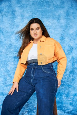 Photo for Body positive and brunette plus size woman with long hair and natural makeup wearing crop top, orange jacket and denim jeans while posing and looking at camera on mottled blue background - Royalty Free Image