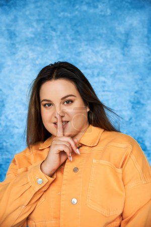 Photo for Body positive and brunette plus size woman with brunette hair and natural makeup wearing orange jacket and showing hush sign and looking at camera on mottled blue background - Royalty Free Image