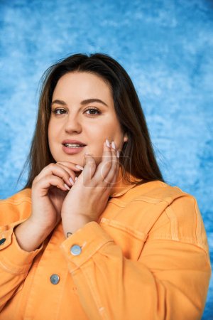 portrait of body positive and brunette plus size woman with long hair and natural makeup wearing orange jacket, touching face and looking at camera on mottled blue background 