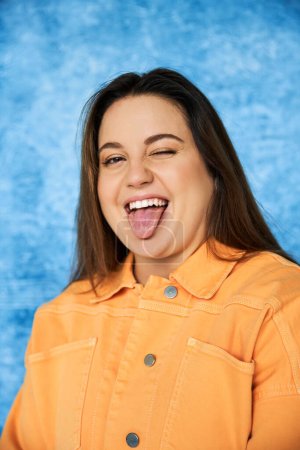 retrato de mujer divertida más tamaño con cabello moreno y maquillaje natural con chaqueta naranja y la lengua sobresaliente mientras mira a la cámara sobre fondo azul moteado, cuerpo positivo 