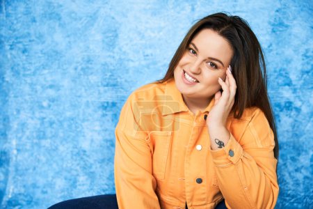 tattooed and smiling plus size woman with brunette hair and natural makeup wearing orange jacket and touching cheek while posing and looking at camera on mottled blue background, body positive 