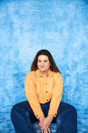 body positive, plus size woman with brunette hair and natural makeup sitting in orange jacket and denim jeans while smiling and looking at camera on mottled blue background 