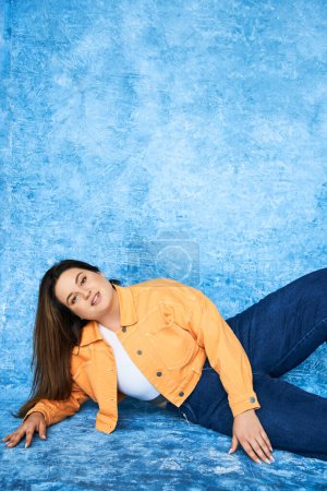 plus size woman with brunette hair and natural makeup wearing crop top, orange jacket and denim jeans while posing and looking at camera on mottled blue background, body positive 