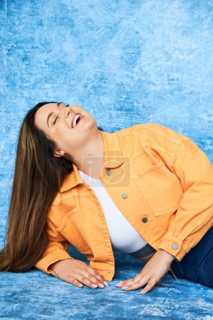 body positive and happy plus size woman with long hair and natural makeup laughing with closed eyes while posing in orange jacket and denim jeans on mottled blue background 