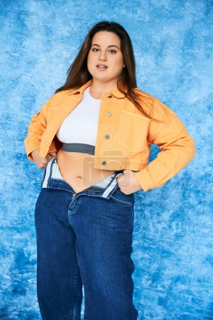 body positive and brunette plus size woman with long hair and natural makeup, in crop top and orange jacket wearing denim jeans while posing and looking at camera on mottled blue background 