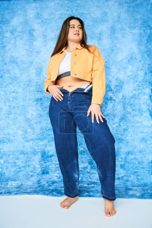 volle Länge der Barfuß-Körper positive Frau mit Plus-Size-Körper und brünetten Haaren posiert in orangefarbener Jacke, Crop Top und Jeans, während posiert und in die Kamera auf fleckigem blauem Hintergrund schaut 