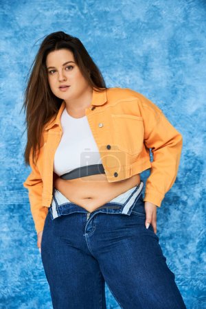 Photo for Body positive and brunette plus size woman with long hair and natural makeup posing in crop top, orange jacket and denim jeans while standing and looking at camera on mottled blue background - Royalty Free Image