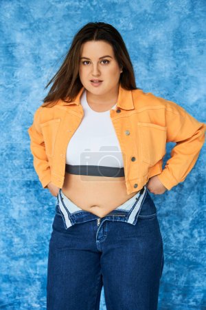 body positive woman with long hair and natural makeup posing in crop top and orange jacket while wearing denim jeans while standing and looking at camera on mottled blue background 