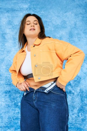 Photo for Brunette body positive woman with natural makeup and closed eyes posing in crop top and orange jacket while wearing denim jeans and standing on mottled blue background, plus size woman - Royalty Free Image