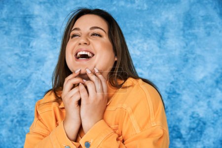 retrato del cuerpo positivo y feliz mujer de talla grande con cabello moreno y maquillaje natural riendo mientras toca la cara y posando en chaqueta naranja sobre fondo azul moteado 