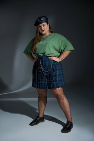full length of plus size woman posing in leather beret, green t-shirt, plaid skirt with chains and fishnet tights looking at camera and posing on dark background, tattoo translation: harmony