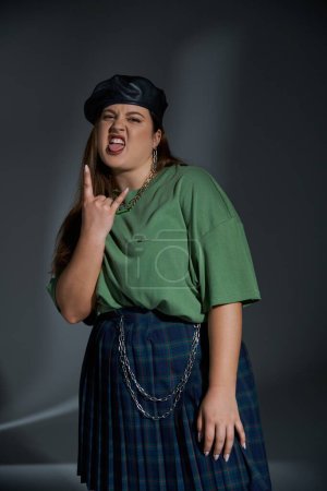stylish plus size woman posing in leather beret, green t-shirt and plaid skirt looking at camera, sticking out tongue and showing rock sign on dark background with studio lighting 