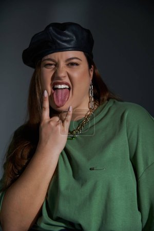 Porträt einer stylischen Plus-Size-Frau, die in Lederbarett und grünem T-Shirt posiert, in die Kamera blickt, während sie die Zunge herausstreckt und Rockzeichen auf dunklem Hintergrund mit Studiobeleuchtung zeigt 