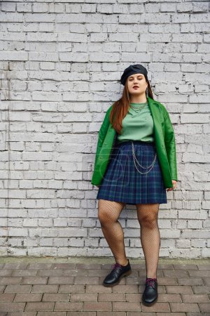 Photo for Full length of chic plus size woman posing in plaid skirt with chains, green leather jacket, beret, fishnet tights and black shoes while posing near brick wall on urban street, body positive - Royalty Free Image