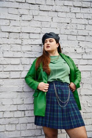 Téléchargez les photos : Chic plus taille femme posant en jupe à carreaux avec des chaînes, veste en cuir vert, béret noir et collants résille tout en regardant loin et debout près du mur de briques sur la rue urbaine, corps positif - en image libre de droit