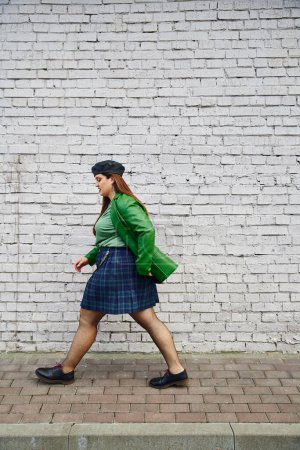 full length of chic plus size woman posing in leather jacket, beret, plaid skirt with chains, fishnet tights and black shoes, walking near brick wall on urban street, body positive 