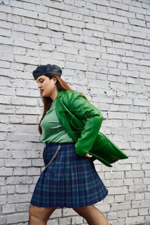 Photo for Side view of stylish plus size woman in green leather jacket, black beret, plaid skirt with chains and fishnet tights walking near brick wall on urban street, body positive, self-love, urban chic - Royalty Free Image