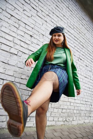Low-Winkelansicht von chic plus size Frau posiert in Leder Baskenmütze und Jacke karierten Rock mit Ketten, Netzstrumpfhosen und schwarzen Schuhen beim Posieren in der Nähe Ziegelmauer auf der städtischen Straße, Körper positiv 