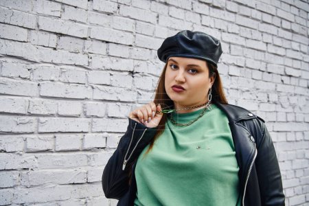 stylish plus size woman in leather jacket and black beret pulling chain necklace while looking at camera near brick wall on urban street, body positive, self-love, unapologetic 