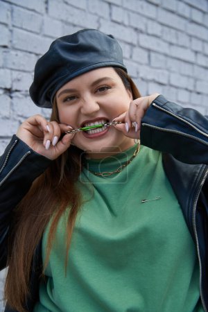 portrait of stylish plus size woman in leather jacket and black beret biting chain necklace while looking at camera and standing near brick wall on urban street, body positive, urban chic