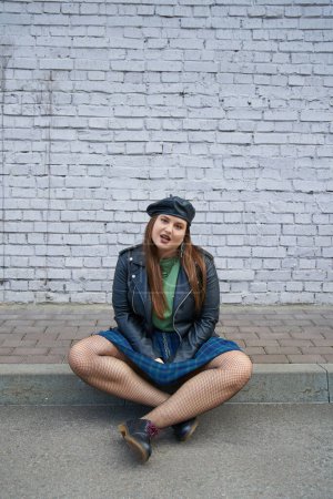 full length of chic plus size woman posing in leather jacket, plaid skirt with chains, fishnet tights and black shoes while sitting near brick wall on urban street, body positive, full length