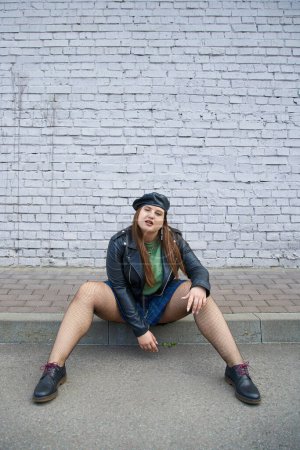 modische Plus-Size-Frau posiert in Lederjacke und Baskenmütze, kariertem Rock, Netzstrumpfhose und schwarzen Schuhen, während sie in der Nähe einer Ziegelmauer auf der städtischen Straße posiert, körperbetont, volle Länge 
