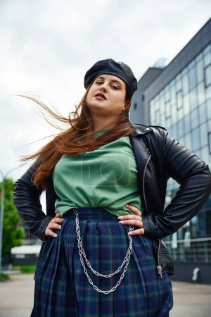 confident woman with plus size body posing in leather jacket with black beret near blurred modern building on urban street outdoors, body positive, hands on hips, looking at camera