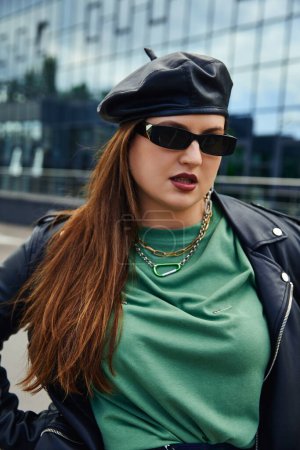 portrait of brunette woman with plus size body posing in stylish sunglasses, leather jacket with black beret, and greet t-shirt near blurred modern building on urban street outdoors, body positive 
