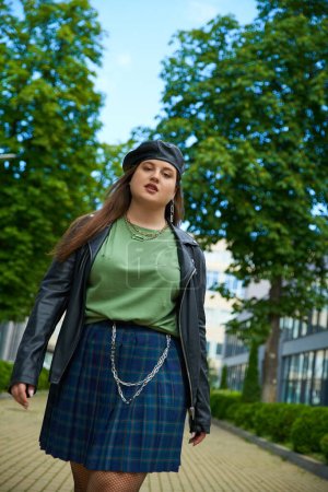 confident plus size woman walking in leather jacket with black beret, plaid skirt and green t-shirt near blurred modern building on urban street outdoors, body positive, trees on background 