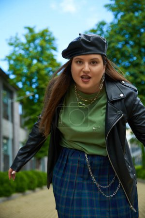 confident woman with plus size body looking at camera and posing in leather jacket with black beret near blurred modern building and green trees on urban street outdoors, body positive 