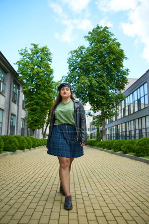 Photo for Full length of plus size woman walking in leather jacket, beret, plaid skirt, fishnet tights and black shoes while looking at camera on urban street with buildings and trees on blurred background - Royalty Free Image